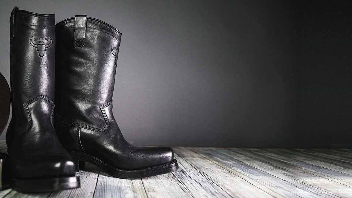 A pair of black Dayton Black Beauty boots on a pale wood floor against a dark grey wall